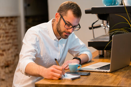 Focused male barista sitting at wooden counter with laptop and taking notes in notebook while working in coffee shop