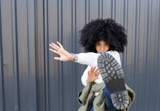 Self assured young ethnic lady with Afro hairstyle in trendy outfit and boots kicking camera while standing on street near metal wall