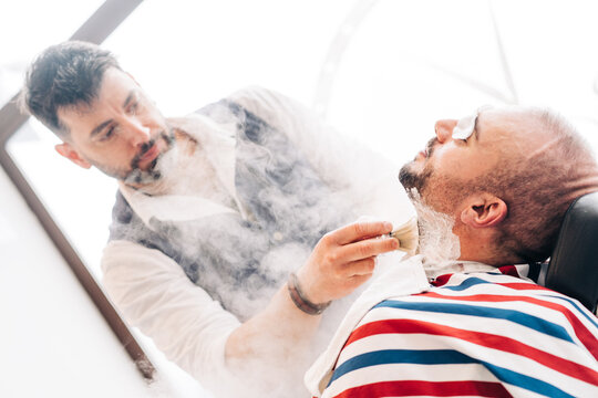 From below male beauty master shaving beard of client with straight razor during steam vapor treatment in hairdressing salon