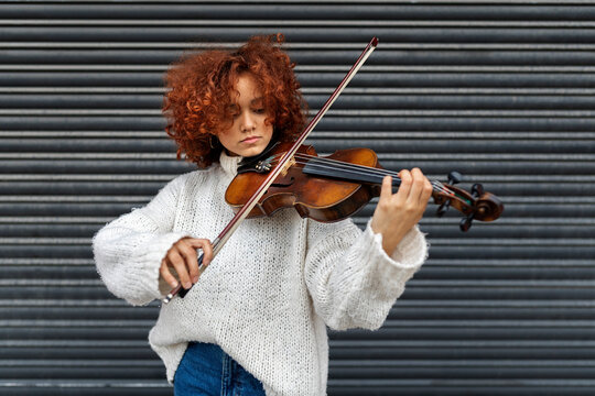 wistful young professional female musician with red curly hair wearing casual sweater standing with acoustic violin against building wall and looking at camera
