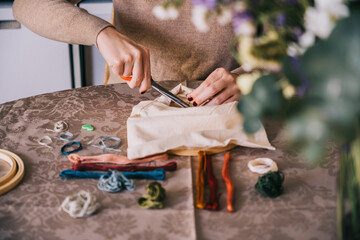 Crop unrecognizable female cutting threads on fabric on hoop while doing embroidery and sitting on table with hoop and threads