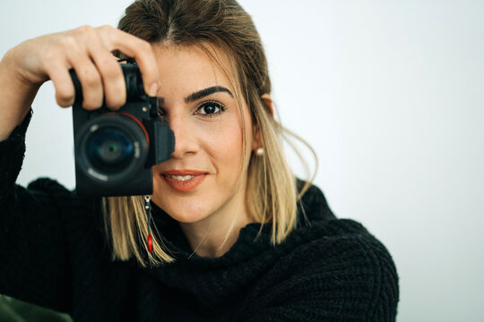 Crop smiling female in black sweater looking at camera while taking photo on professional digital camera in house on white background
