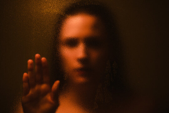 Anonymous blurred female touching glass while standing in dark room with orange illumination