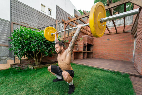 Muscular male with strong naked torso doing clean and jerk weightlifting exercise with barbell during training in courtyard in summer