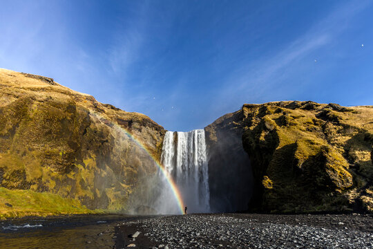 Unrecognizable traveler standing near amazing waterfall and rainbow in mountains during vacation in Iceland