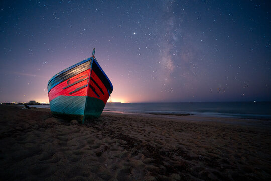Low angle of wooden boat moored on sandy shore near sea on background of night starry sky in long exposure in Cadiz