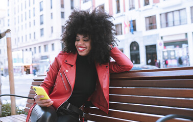 Stylish astonished African American female reading news on cellphone sitting on bench in city