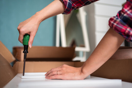 Crop faceless female sitting on floor and screwing screws in wooden board while assembling new furniture at home