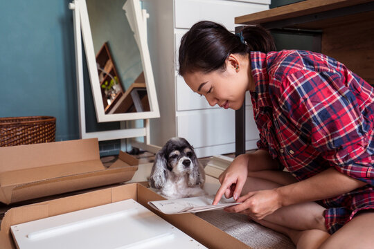 Smiling Asian female sitting on floor with fluffy dog and reading instruction while assembling furniture in new apartment