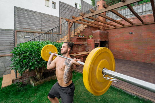 Muscular male with strong naked torso doing clean and jerk weightlifting exercise with barbell during training in courtyard in summer
