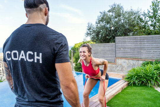 Cheerful female athlete doing resistance band bent over rows during workout under supervision of personal trainer