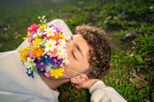 From Above Of Anonymous Young Male With Colorful Blossoming Flower Mask On Face And Closed Eyes Lying On Lawn In Sunlight