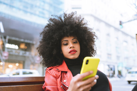 Stylish astonished African American female reading news on cellphone sitting on bench in city