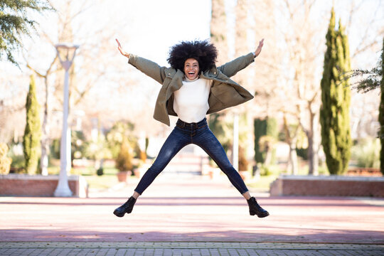 Full length of cheerful young ethnic woman with Afro hair in stylish outfit yelling and jumping in park on sunny day