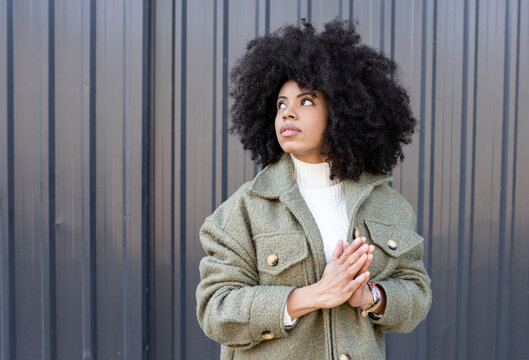 Pensive young stylish African American female millennial with curly hair in trendy outfit standing on street near metal fence and looking away