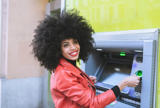Side view of positive African American female standing near ATM and inserting credit card while smiling widely and looking away