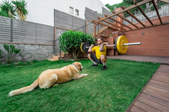 Athletic male in sportswear doing clean and jerk exercise with barbell during workout in backyard with fluffy dog