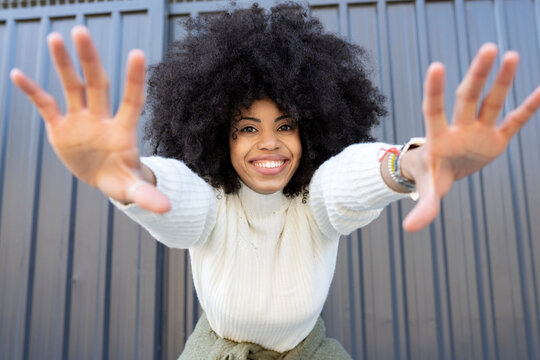 Cheerful young African American lady with curly hair in casual clothes showing two hands and looking at camera on street