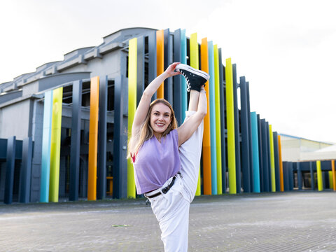 Full length attractive fit female in casual wear performing standing split and looking at camera with smile while standing on against creative building on sunny summer day