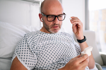 Crop adult male patient in hospital gown putting on TWS earbuds while lying on bed in modern ward in clinic