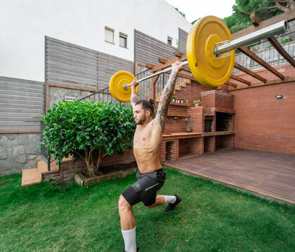 Muscular male with strong naked torso doing clean and jerk weightlifting exercise with barbell during training in courtyard in summer