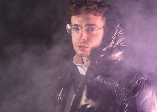 Thoughtful young unshaven guy in trendy outfit against black background and smoke