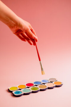 Crop unrecognizable creative person dipping paintbrush on colorful palette of watercolor paints against pink background