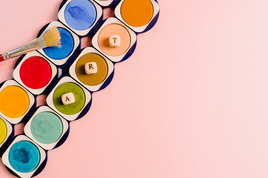 Top view composition with paintbrush placed near colorful palette of watercolor paints with small cubes forming word Art on pink background