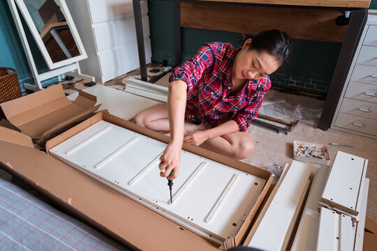 From above ethnic Asian female sitting on floor and screwing screws in wooden board while assembling new furniture at home