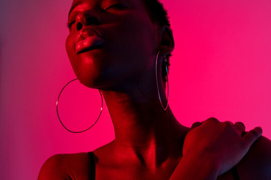 Self assured young African American female model in black top with closed eyes while standing against pink background in neon light