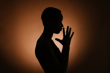Silhouette of anonymous ethnic female clasping hands and praying against brown background