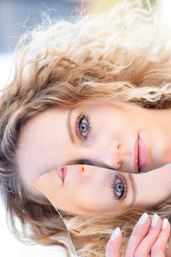 Emotionless female with blond curly hair covering eye with piece of broken mirror with reflection of face and looking at camera