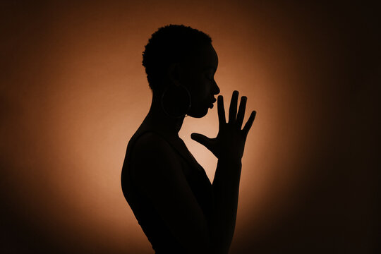 Silhouette of anonymous ethnic female clasping hands and praying against brown background