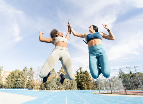 From below of delighted multiethnic female athletes in sportswear in moment of jumping above stadium track giving high five and looking at each other