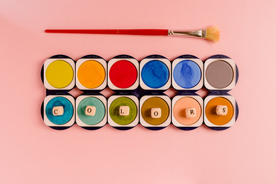 Top view composition with paintbrush placed near colorful palette of watercolor paints with small cubes forming word Colours on pink background