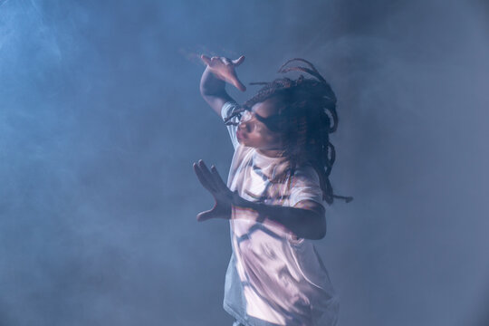 Dynamic African American teenage girl making movement while performing urban dance in neon light against blue background and smoke