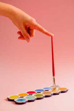 Crop unrecognizable creative person dipping paintbrush on colorful palette of watercolor paints against pink background