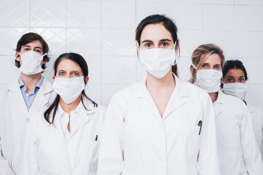 A Group Of Five Doctors With A Mask On Their Face And A White Coat Standing Up And Facing The Camera In A Hospital Room