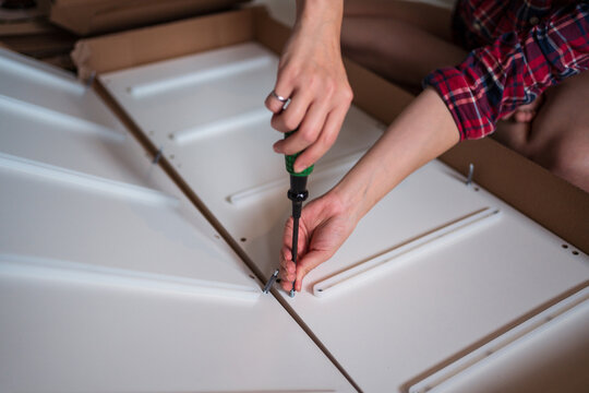 Crop faceless female sitting on floor and screwing screws in wooden board while assembling new furniture at home