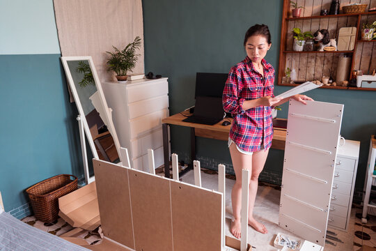 High angle of busy Asian female in domestic clothes standing with instruction and assembling furniture in new flat