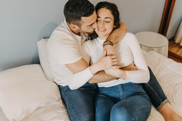 Loving couple sitting on soft bed in bedroom and tenderly hugging while enjoying weekend together at home