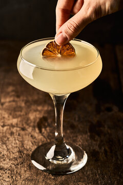 High angle of crop anonymous person adding dried orange slice in glass with yellow alcohol cocktail placed on table