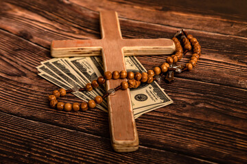 A Catholic cross, a rosary with beads and dollars lie on a dark brown wooden table.