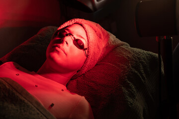 From above of crop anonymous female cosmetologist adjusting protective goggles of relaxed young woman lying on couch during red LED light facial therapy in professional salon