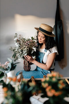Young female horticulturist in straw hat creating bouquet on table with assorted tools at home