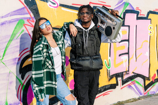 Young black hipster man holding cassette and standing near female partner in modern sunglasses on street in sunlight