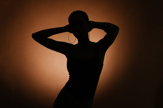 Silhouette of unrecognizable slender female with hands behind head dancing against brown background