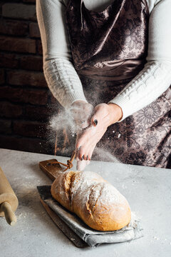 Crop anonymous female artisan in apron sprinkling delicious loaf of white bread with flour at table in bakehouse