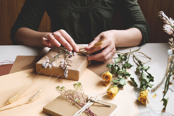 Woman makes zero waste, plastic free, trendy hand made gift package with craft recycled paper and dried flowers on the table with linen tablecloth. Natural aestetic.