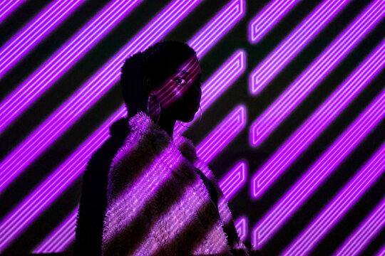 African American woman in stylish fur coat looking at camera while standing under violet neon stripes